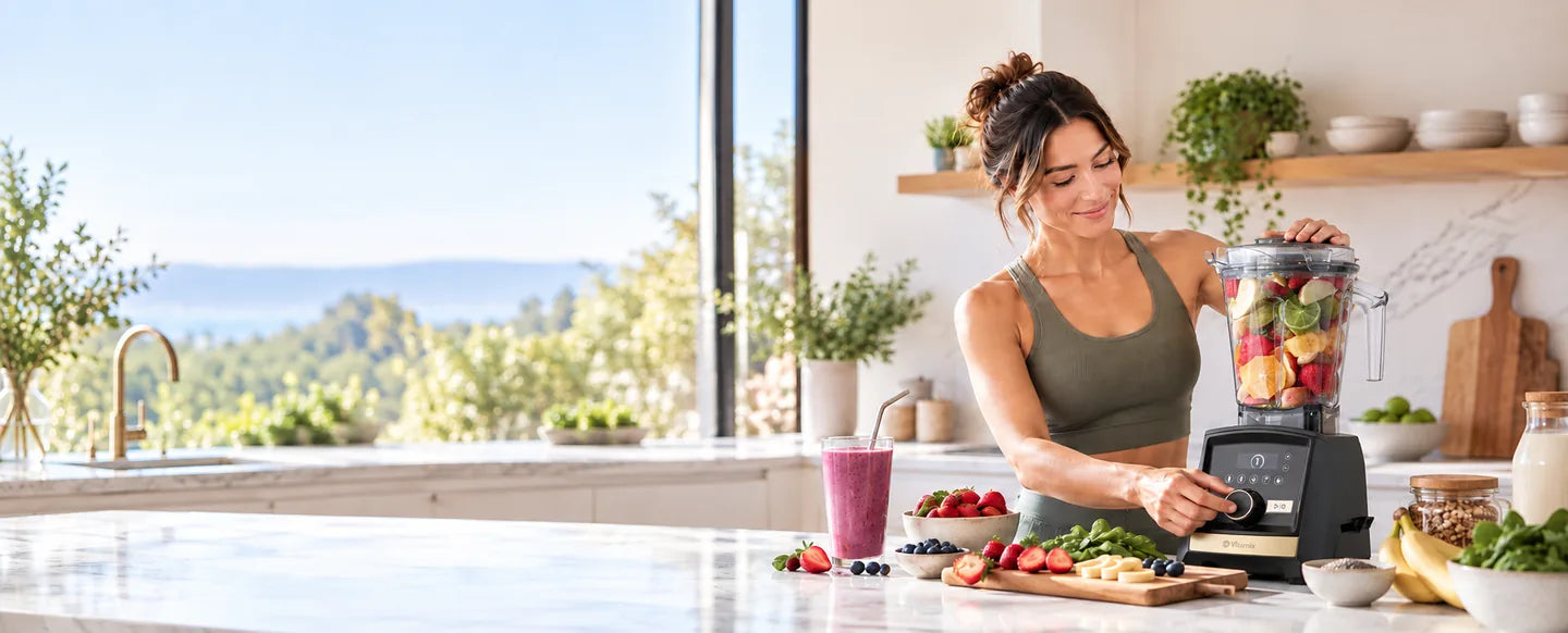 A smiling woman making a fruit smoothie with a Vitamix blender in a sunny kitchen with a coastal view