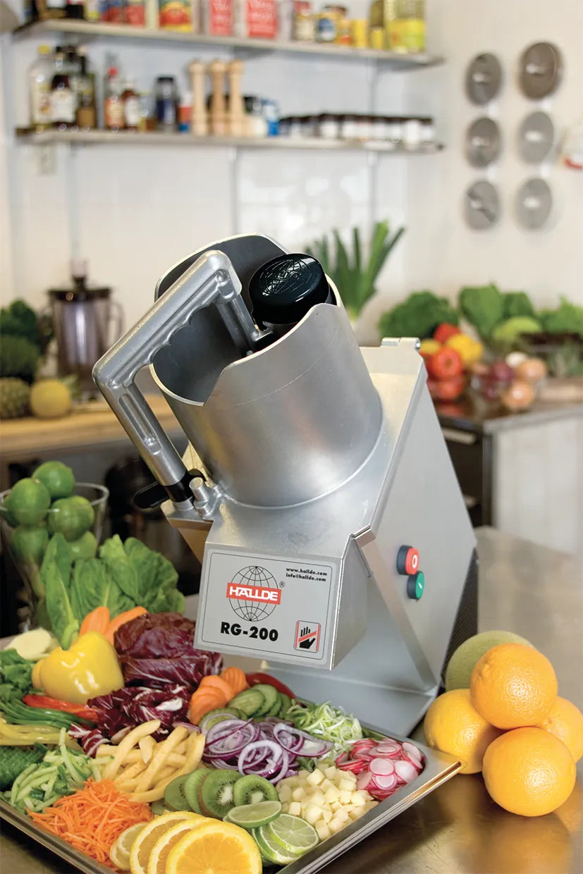 A Hallde RG-200 vegetable preparation machine on a kitchen bench beside a tray of freshly cut vegetables and fruit