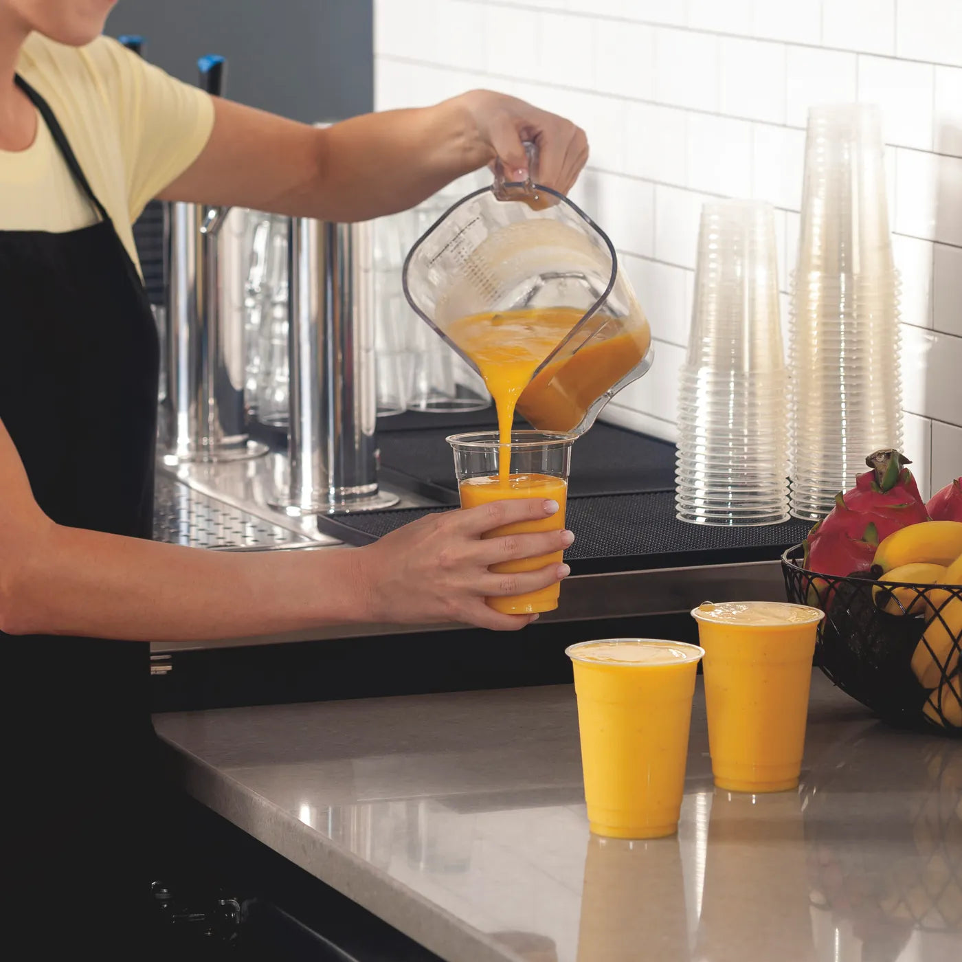 Pouring a mango smoothie from a Hamilton Beach jug into takeaway cups on a cafe bench