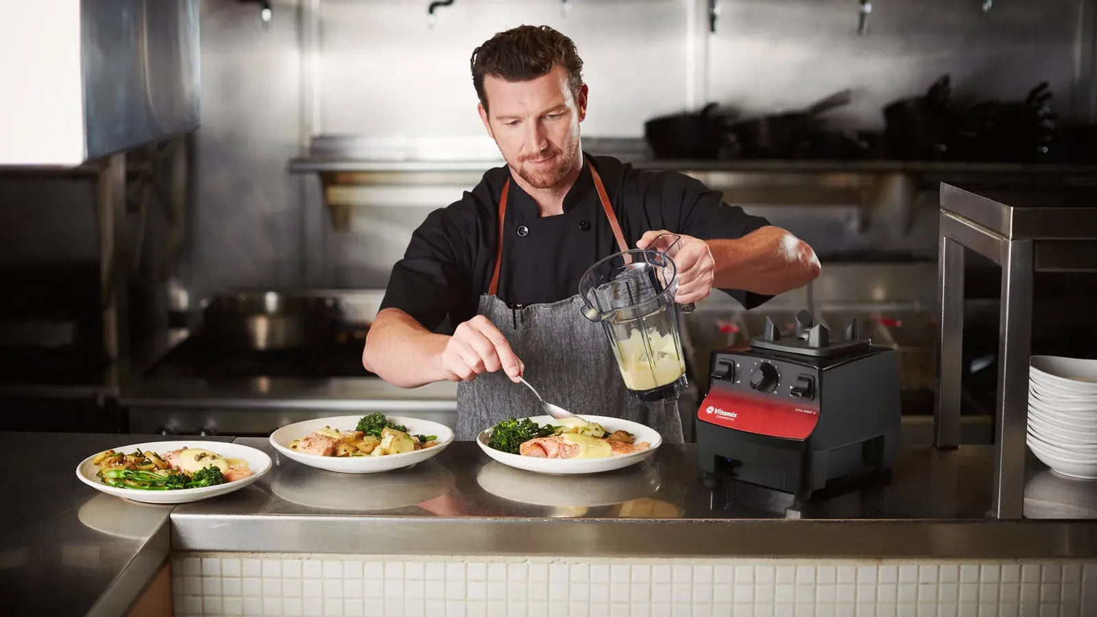 A chef saucing plates from a Vitamix jug, a red Vita-Prep 3 on the stainless pass